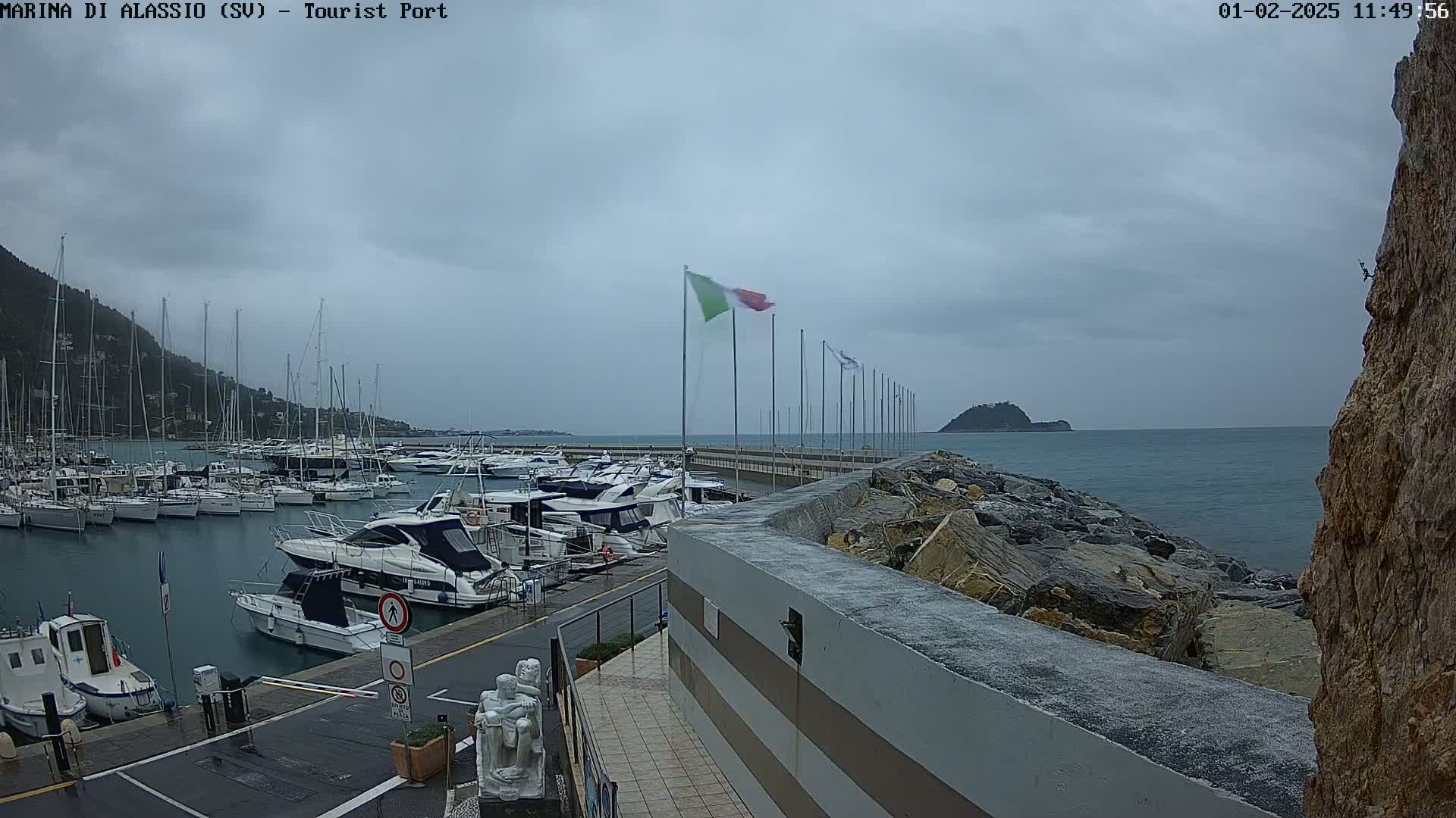 A marina filled with sailboats and motorboats is shown on an overcast day, with a rocky seawall and a small island visible in the distance.