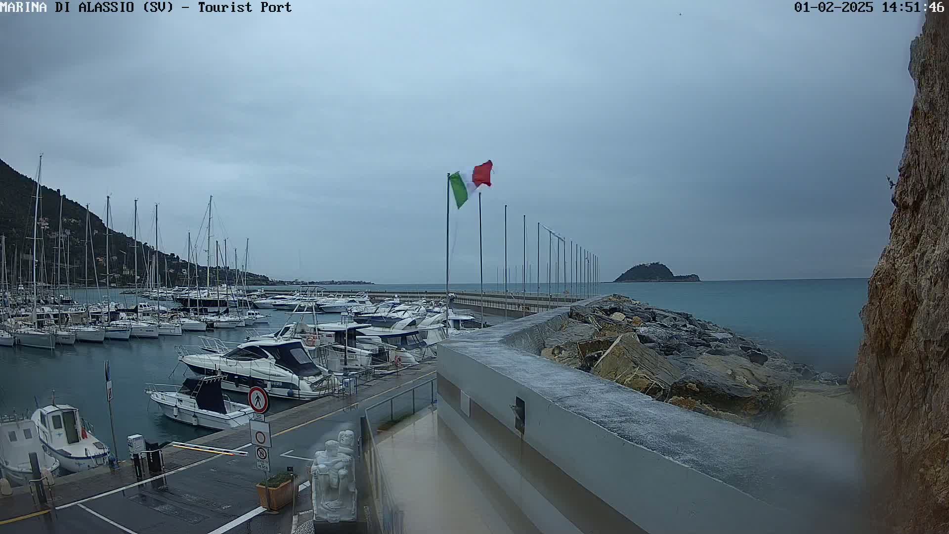 A harbor filled with numerous boats is seen on an overcast day, with a partially visible Italian flag waving in the wind and a rocky shore in the foreground.