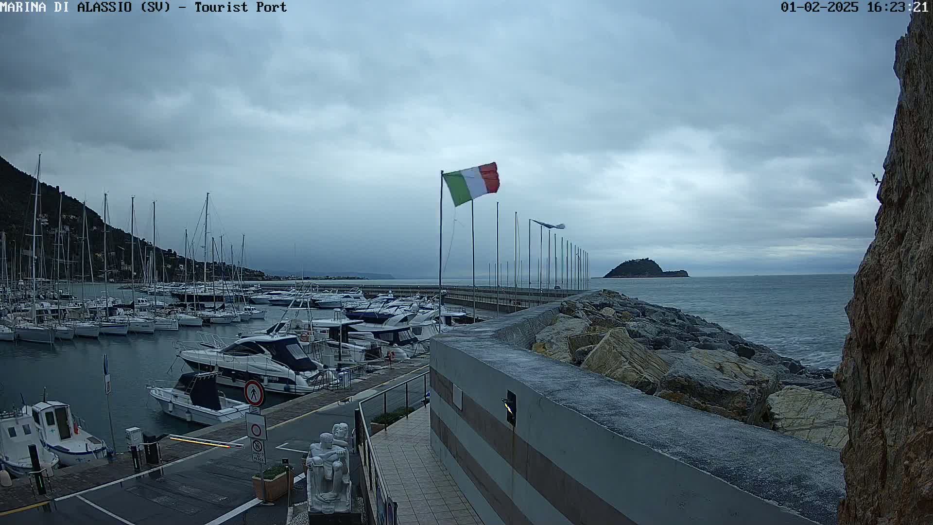 A marina filled with sailboats and motorboats is shown under an overcast sky, with a rocky coastline and a small island visible in the distance.