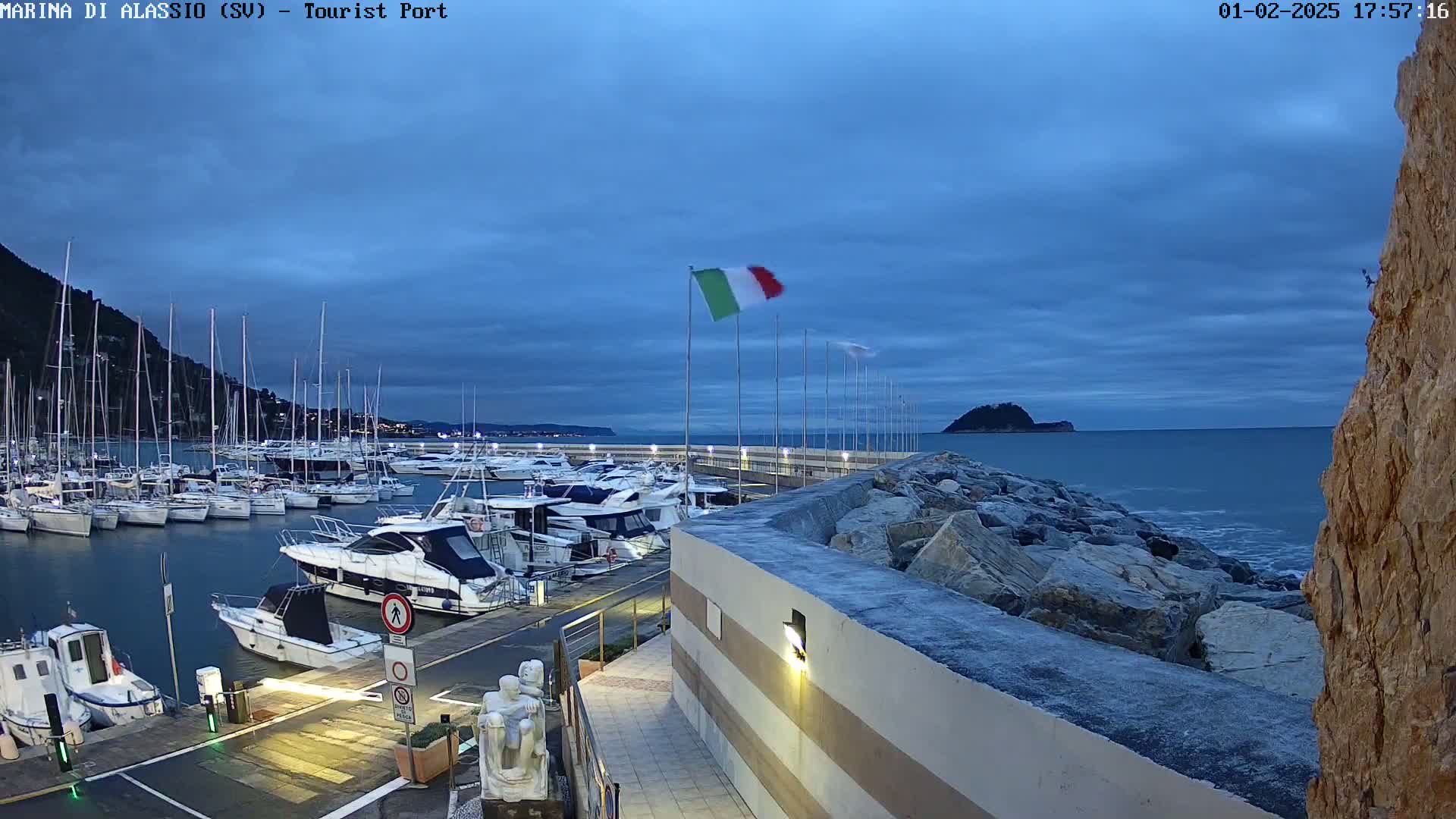 A marina filled with boats is situated on a calm sea under a twilight sky, with a rocky breakwater and an Italian flag waving in the breeze.