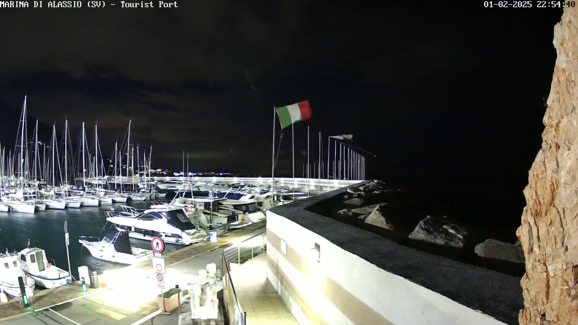 A nighttime view of a marina filled with sailboats and motorboats, under a dark sky with an Italian flag waving in the wind.