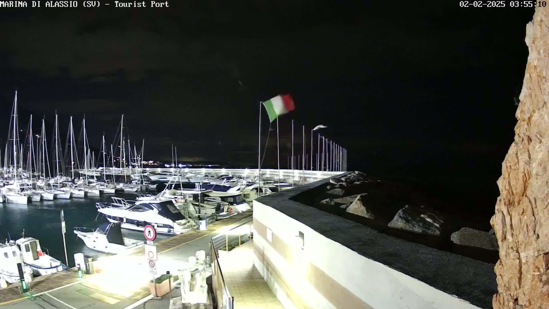 A nighttime view of a marina filled with sailboats and motorboats under a dark sky, with an Italian flag waving in the wind.