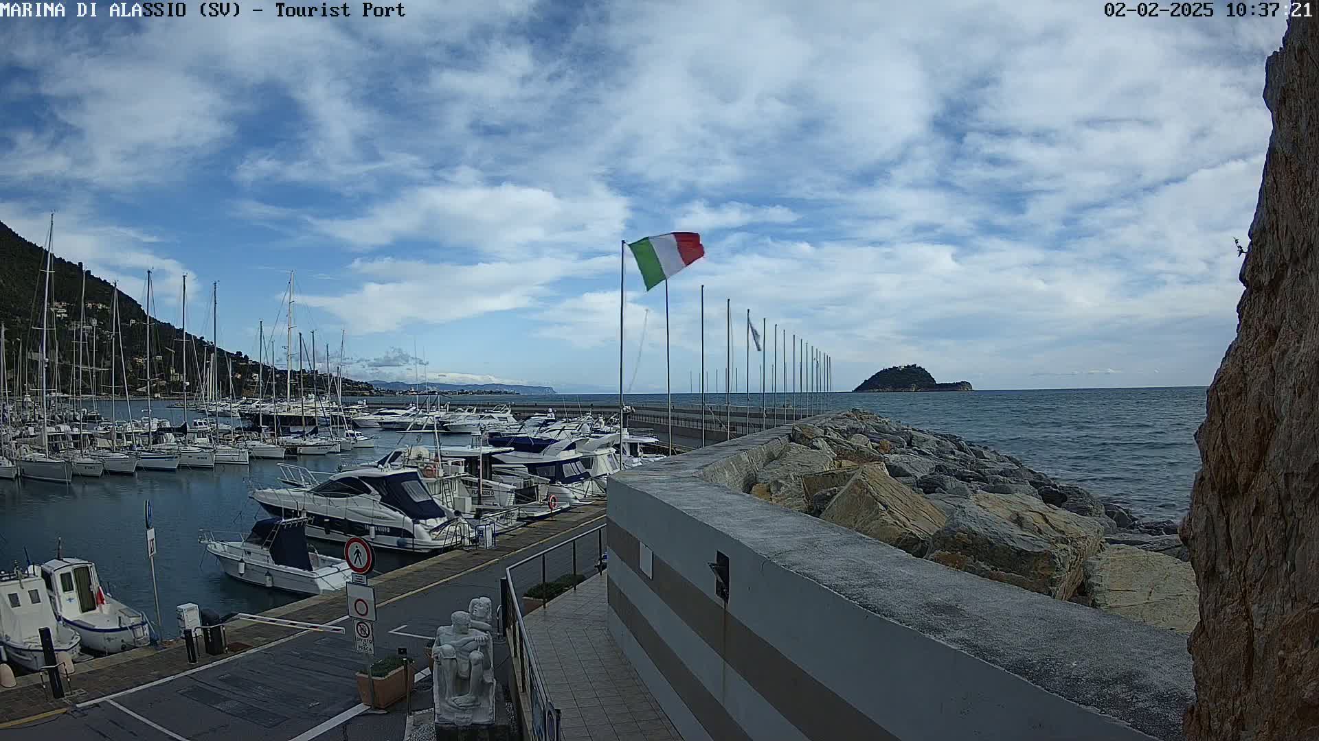 A marina filled with boats is situated on a partly cloudy day next to a rocky shoreline and a small island in the distance.