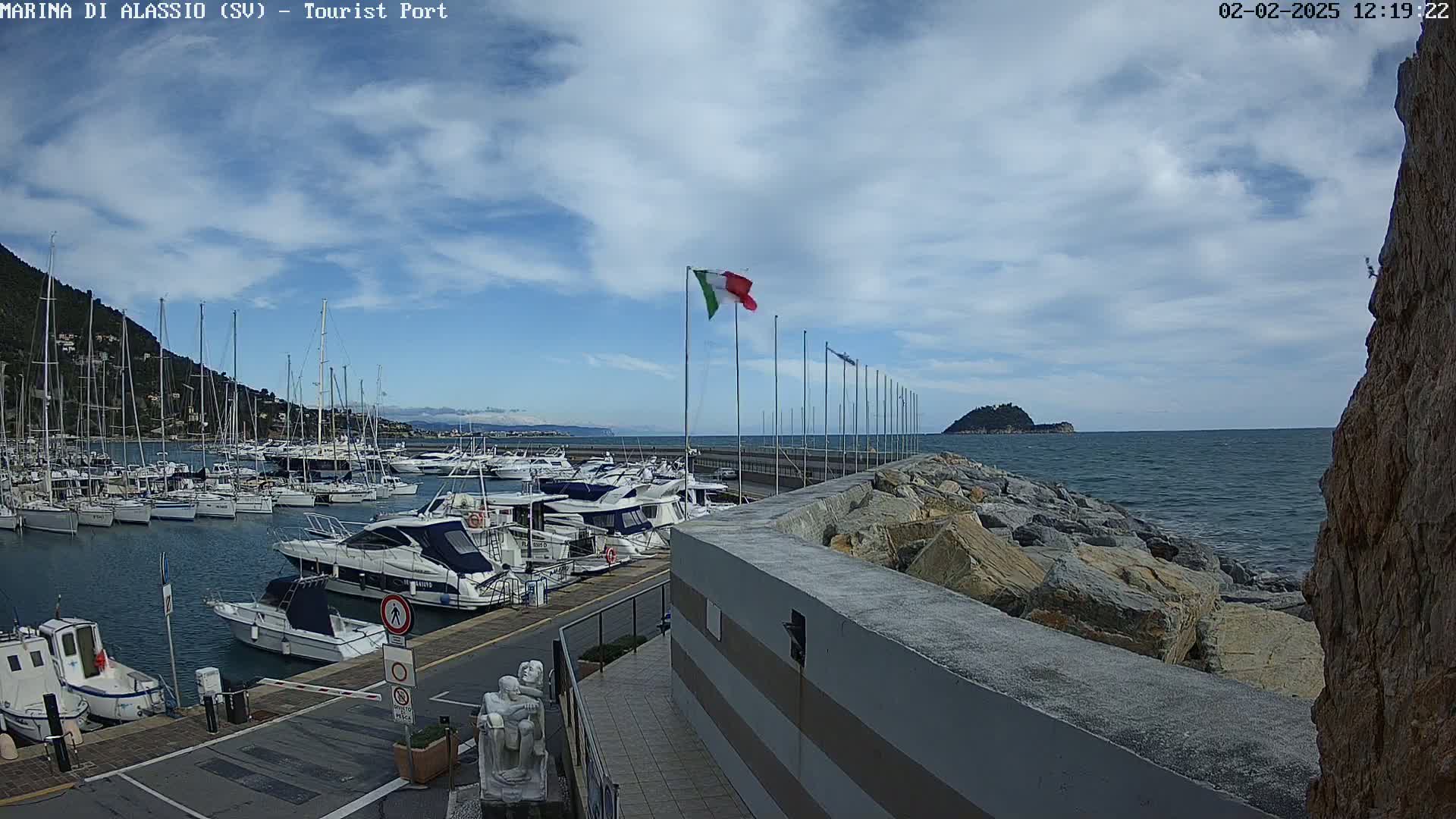 A marina filled with boats is situated next to a rocky shoreline under a partly cloudy sky.