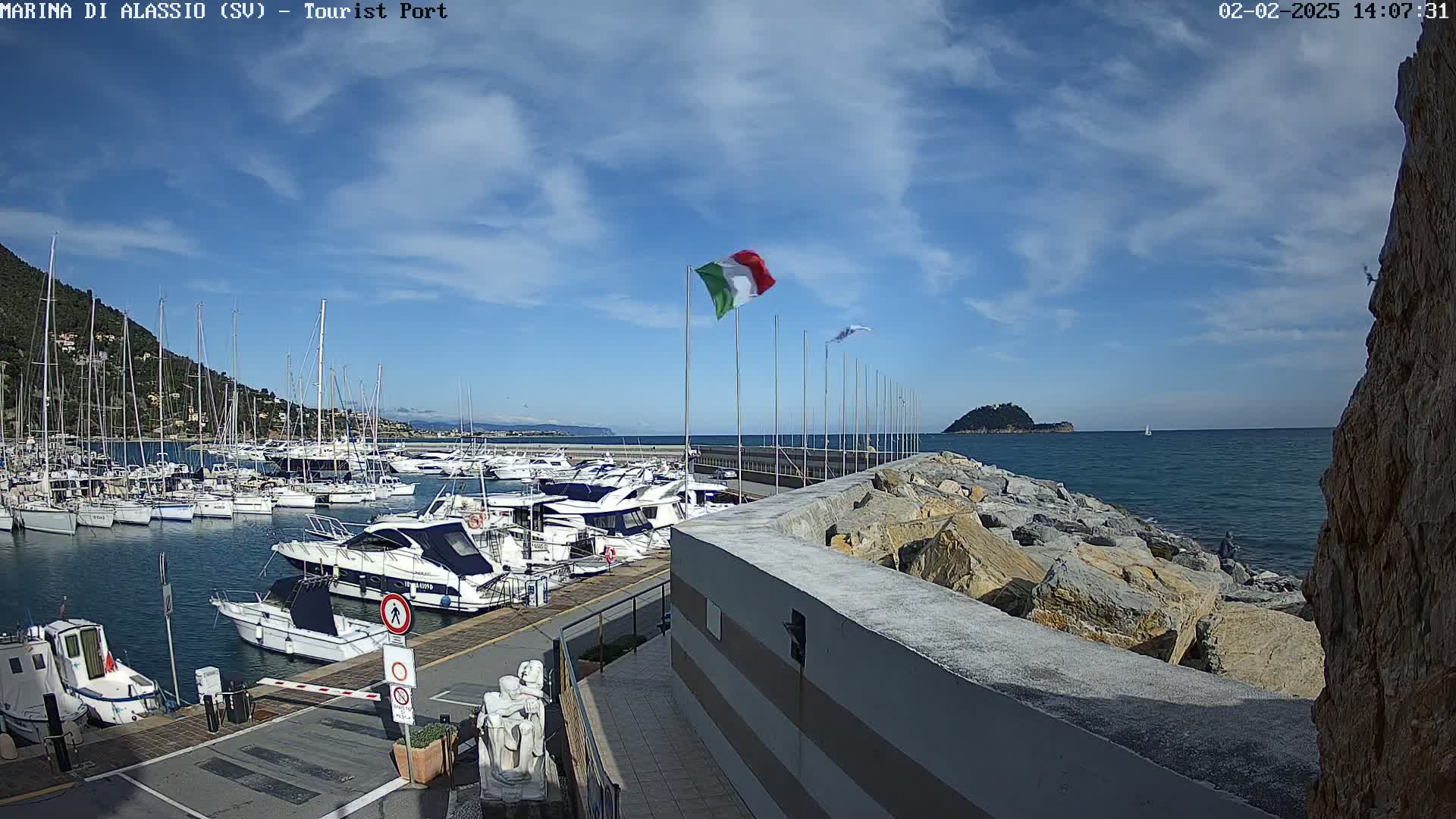 A marina filled with sailboats and motorboats is situated on a calm, sunny day next to a rocky shore with an Italian flag waving in the breeze.