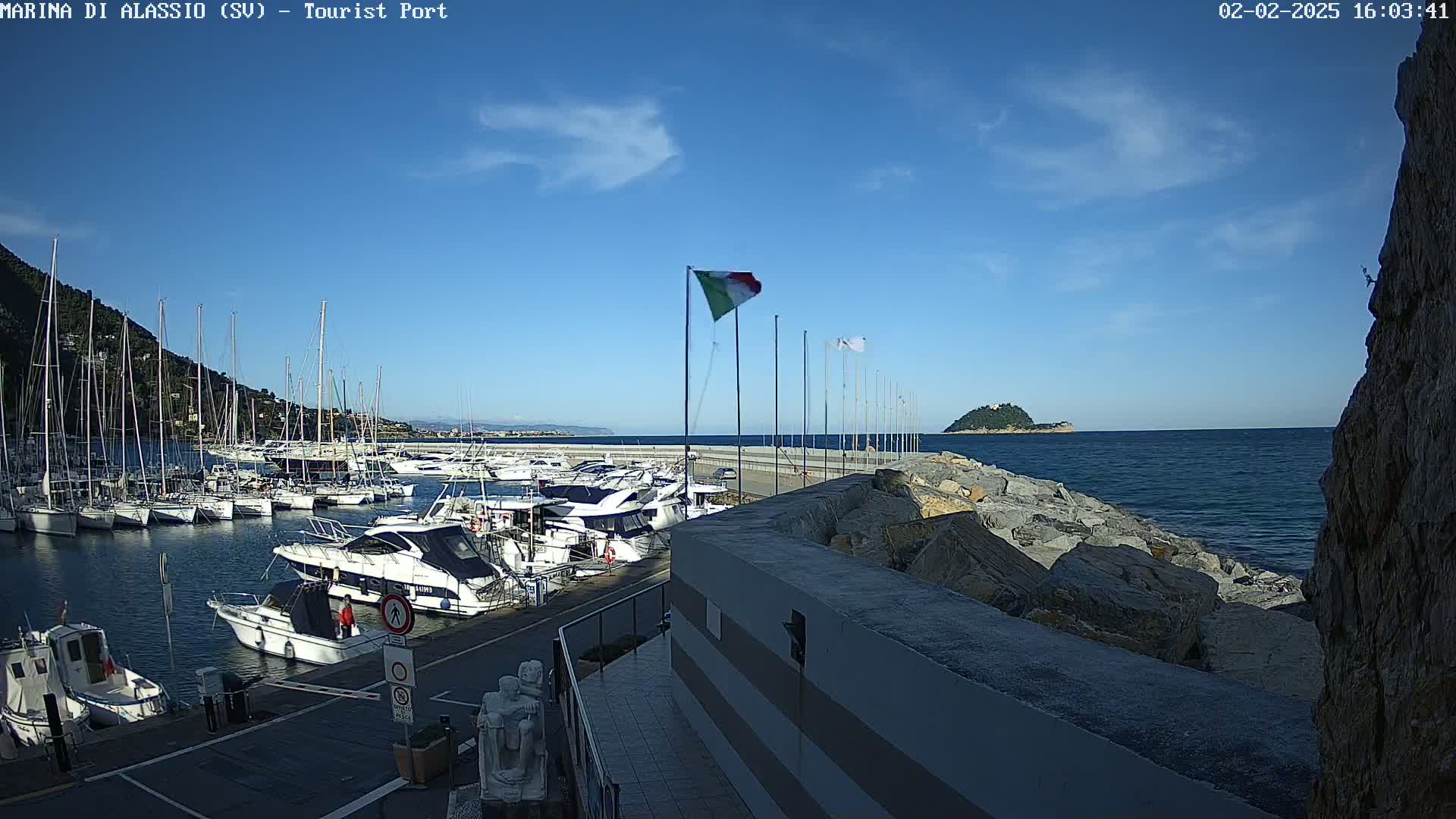 A harbor filled with sailboats and motorboats is situated next to a rocky coast under a clear blue sky.