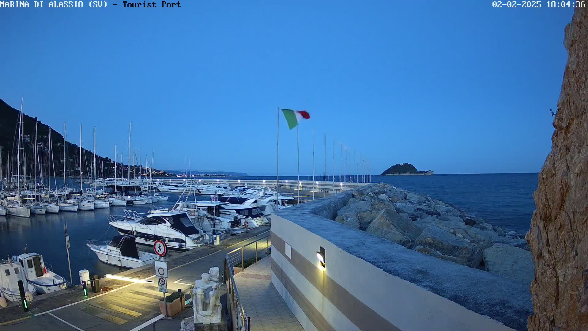 A marina filled with yachts and sailboats is situated on a calm, twilight sea next to a rocky coast, with an Italian flag waving in the breeze.