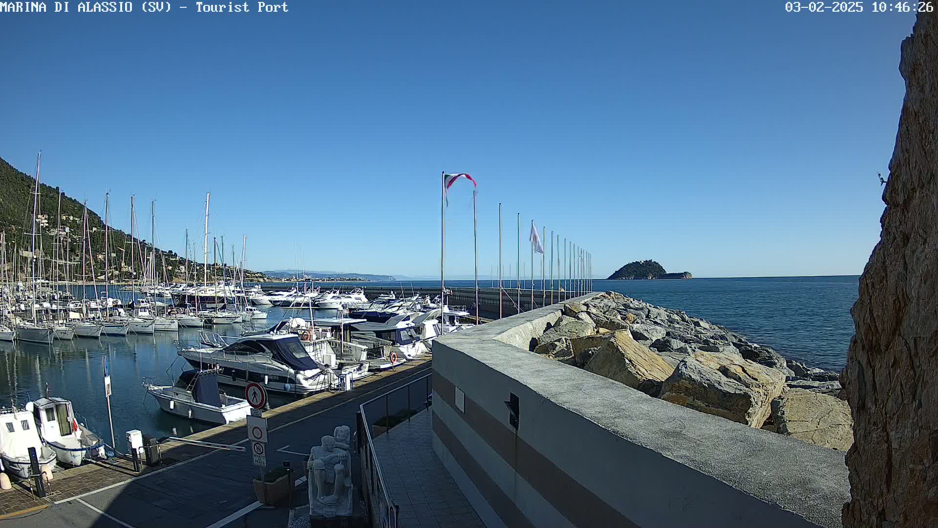 A marina filled with sailboats and motorboats is situated on a calm, clear day next to a rocky seawall and a small island in the distance.