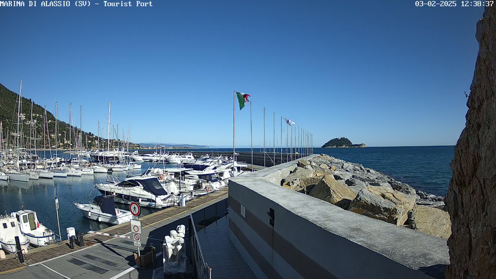 A marina filled with sailboats and motorboats is shown on a sunny day, next to a rocky breakwater with a small island visible in the distance.