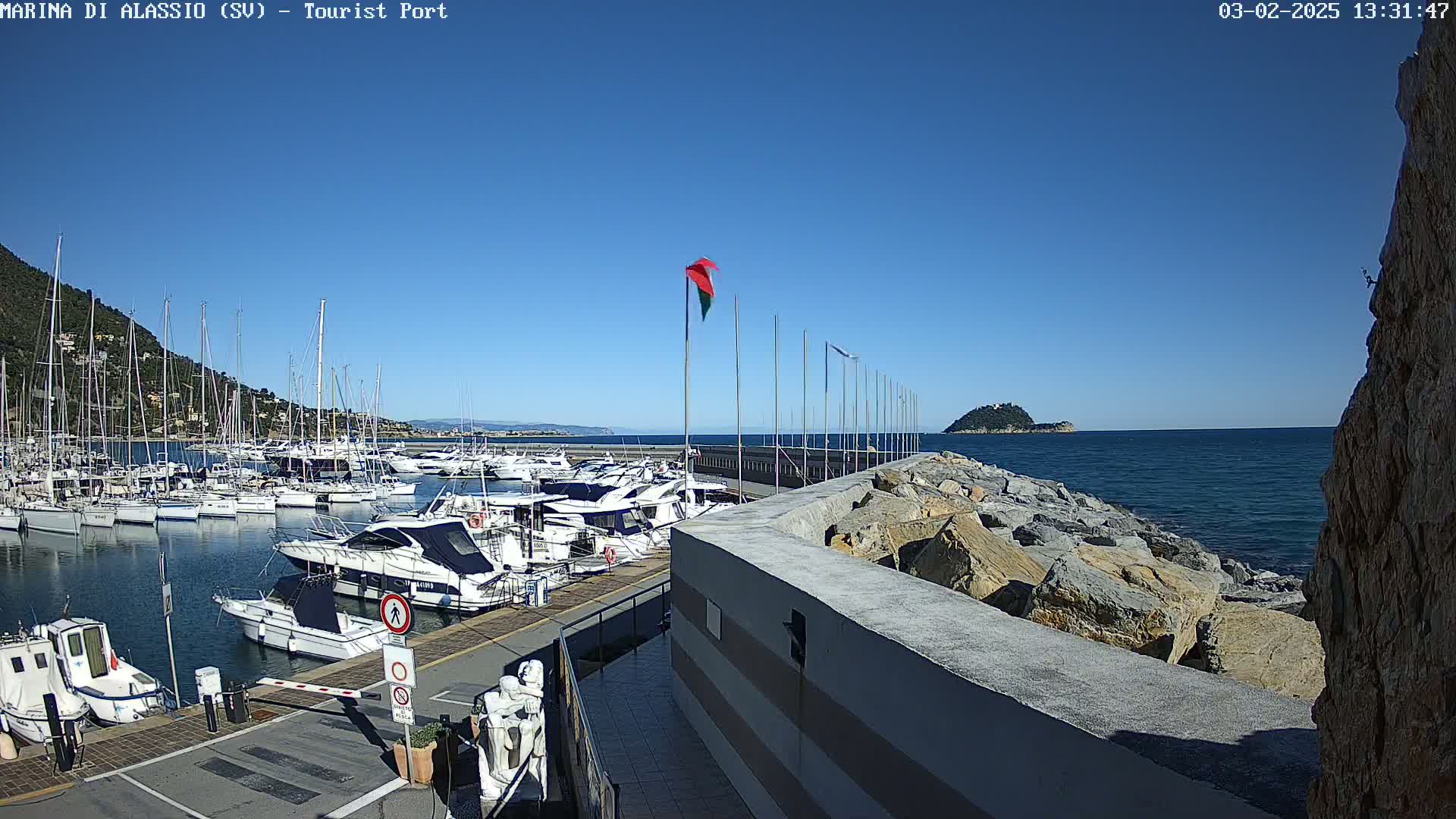 A marina filled with numerous boats is situated on a calm, clear day next to a rocky shoreline and a small island in the distance.