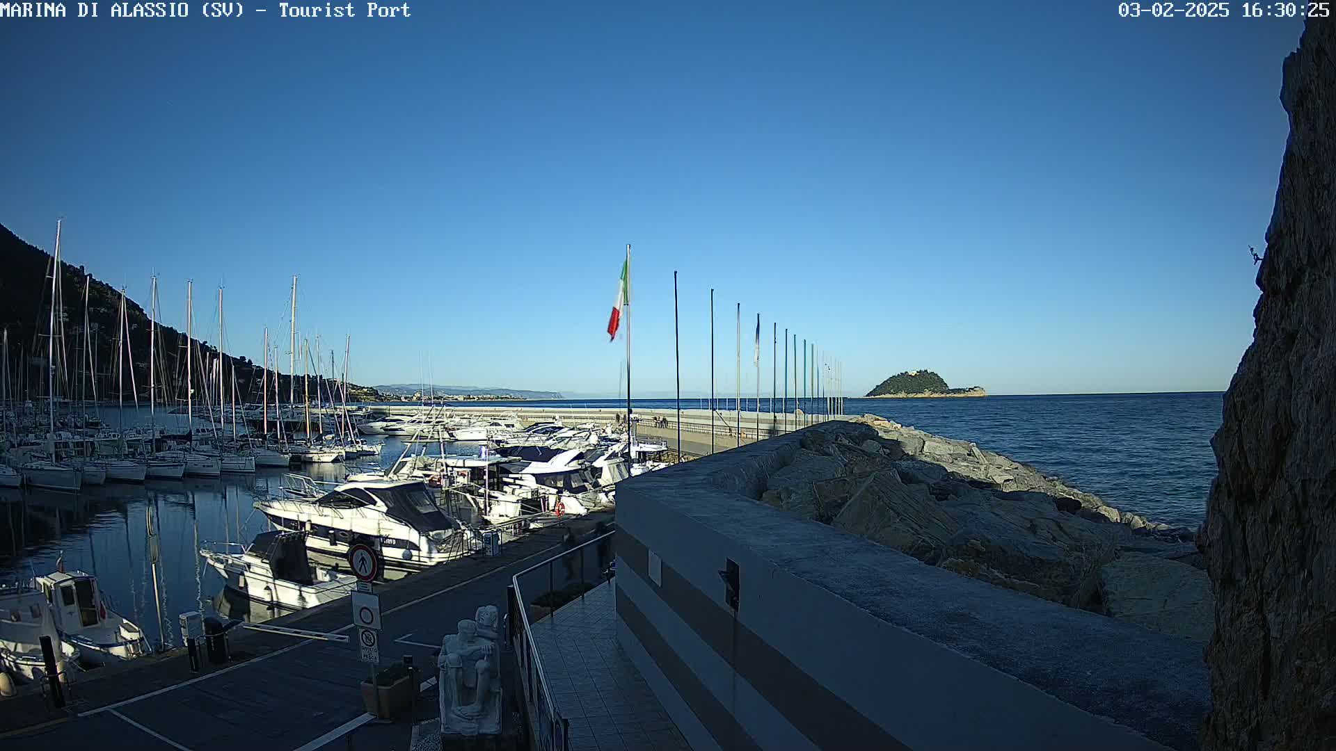 A marina filled with sailboats and motorboats is situated next to a rocky shoreline on a clear, sunny day.