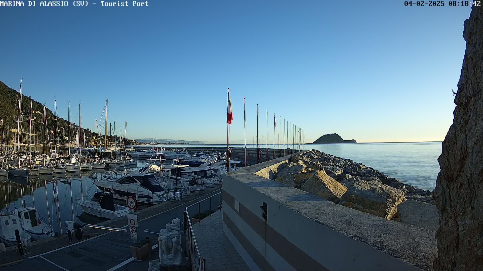 A marina filled with sailboats and motorboats is situated on a calm, clear day next to a rocky breakwater and a small island in the distance.