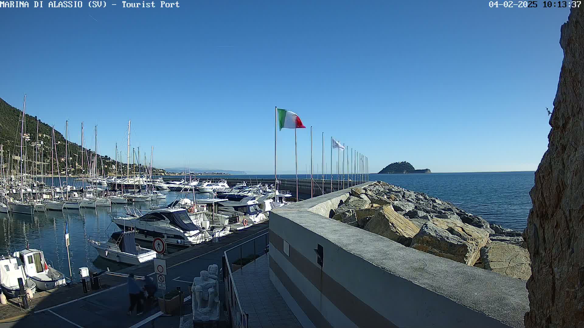 A marina filled with boats sits on a calm, clear day next to a rocky shore and a small island in the distance.