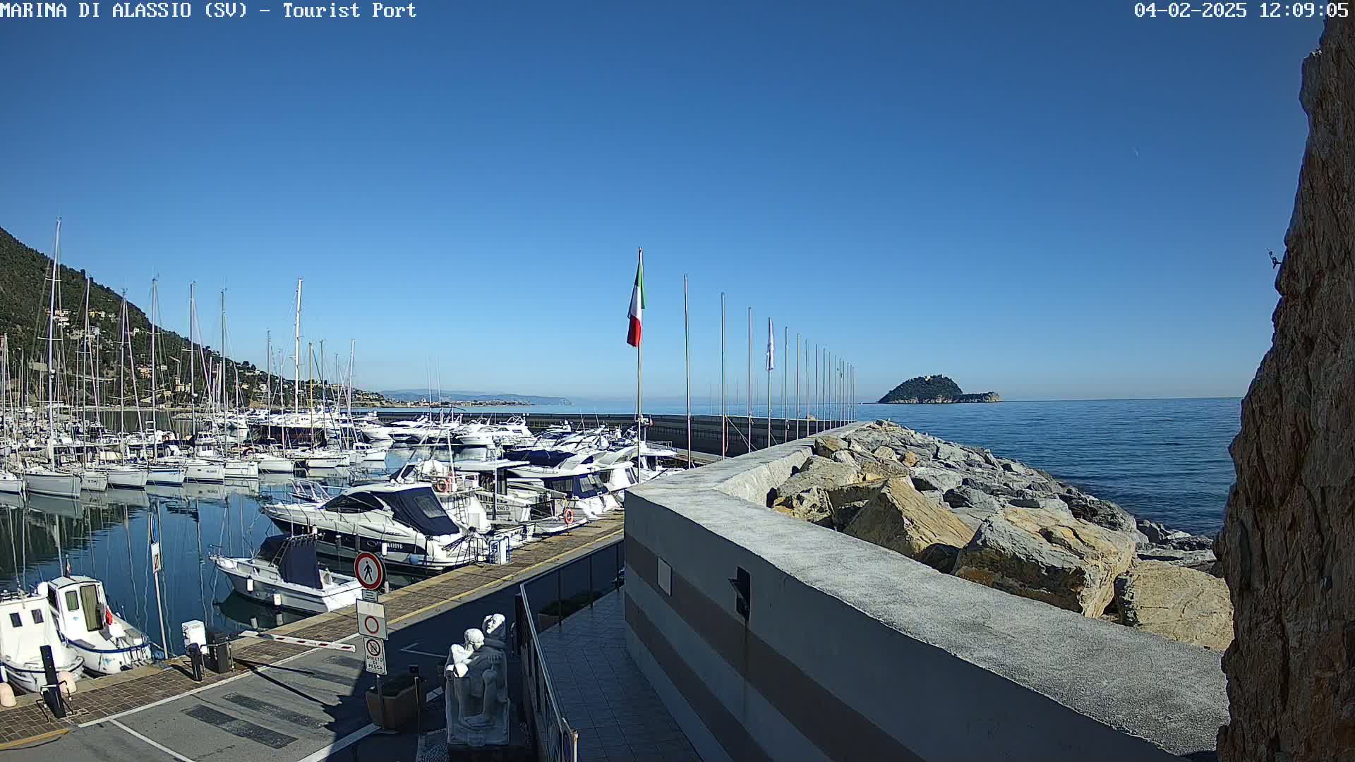 A marina filled with many boats is situated next to a rocky shoreline on a sunny day with a clear blue sky.