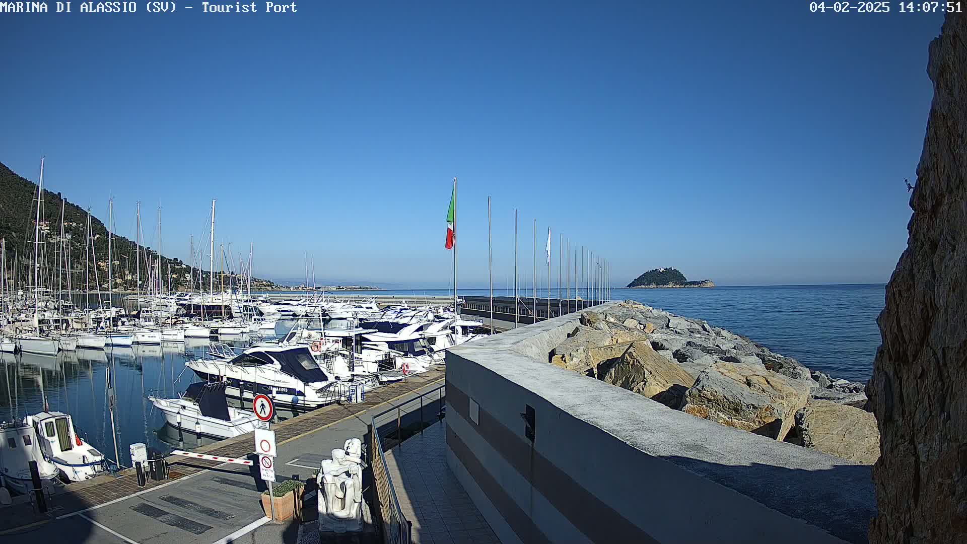 A marina filled with numerous boats is situated next to a rocky shoreline on a clear, sunny day, with a small island visible in the distance.