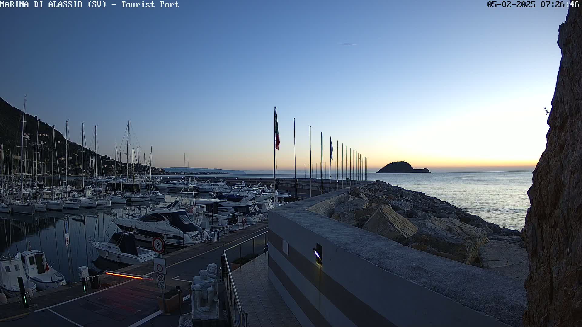 A marina filled with boats is seen at sunrise, next to a rocky shore and calm sea.