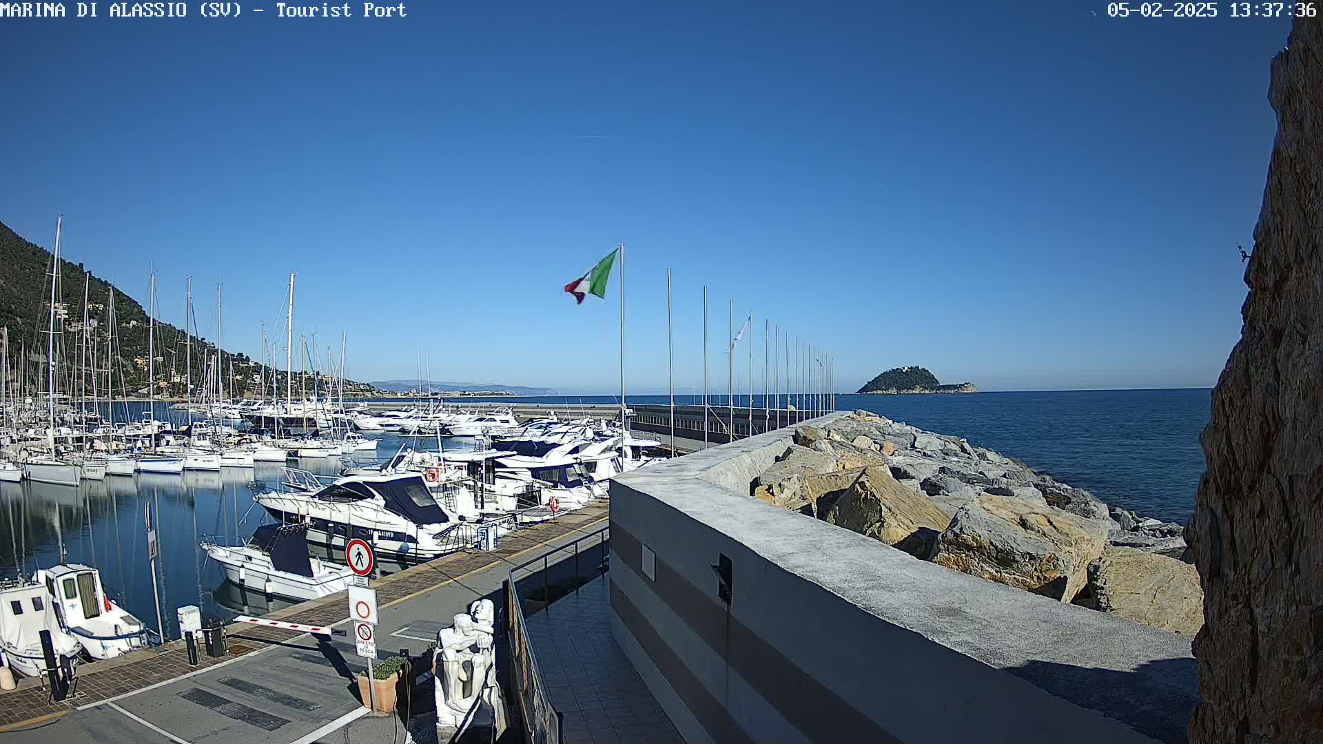 A marina filled with sailboats and motorboats is situated on a calm, sunny day next to a rocky shoreline and a small island in the distance.