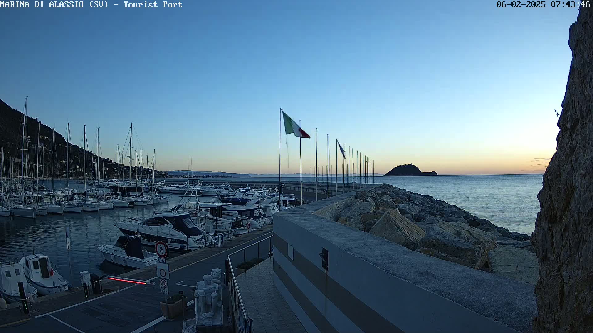 A marina filled with boats is seen at dawn under a clear blue sky, with a rocky breakwater separating it from the calm sea and a small island visible in the distance.