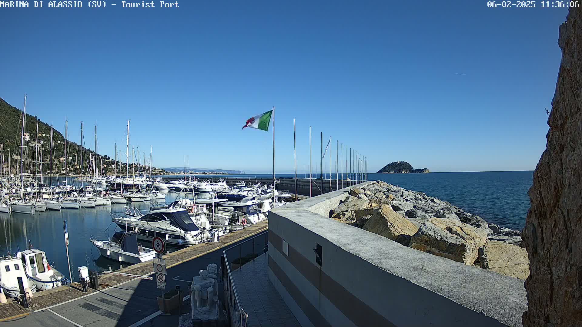 A marina filled with boats is situated next to a rocky seawall on a clear, sunny day.