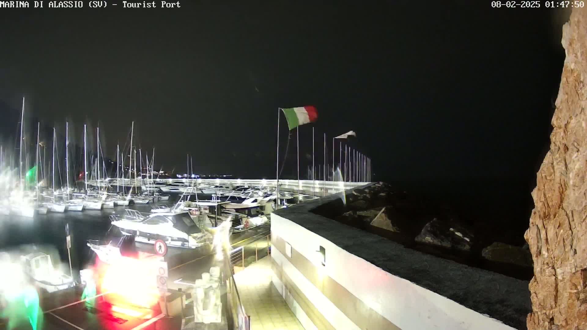 A nighttime view of a marina filled with boats, with an Italian flag waving in the wind and a rocky shoreline visible.