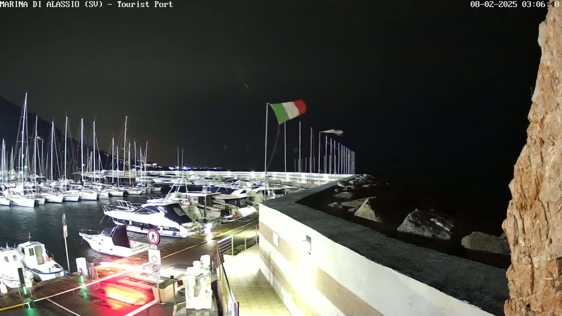 A marina filled with sailboats and motorboats at night under a dark, windy sky, with an Italian flag waving.