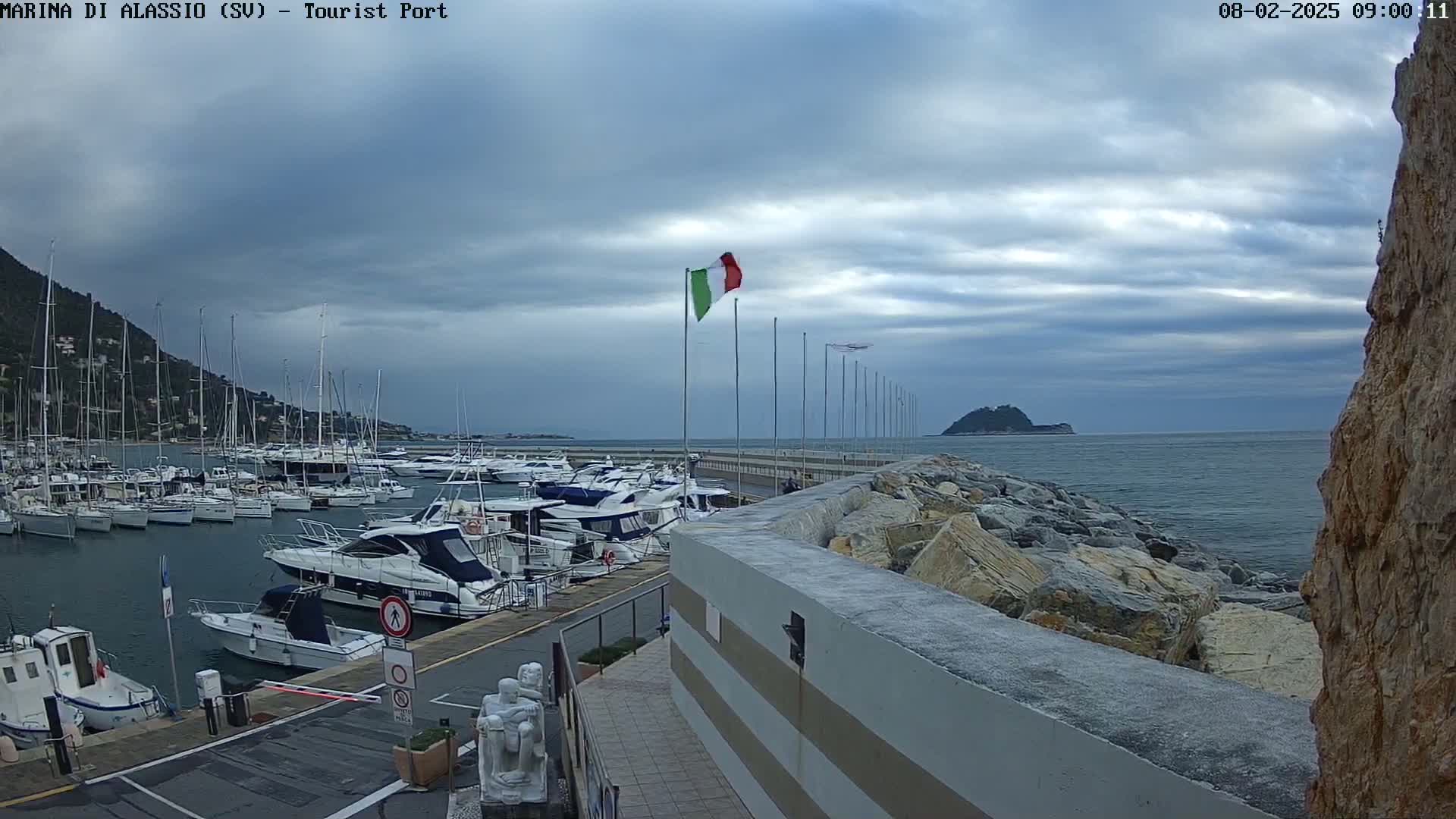 A harbor filled with sailboats and motorboats is situated next to a rocky breakwater under a cloudy sky.