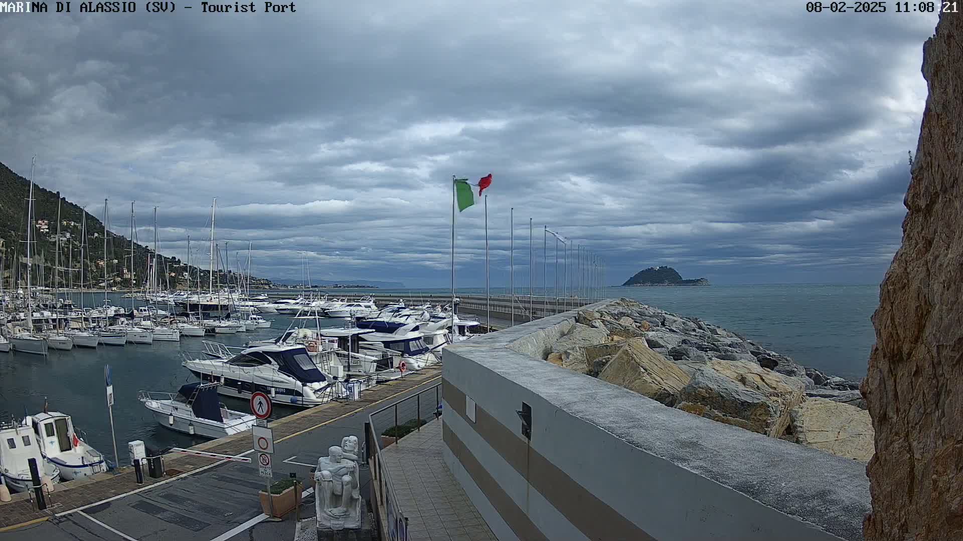 A marina filled with numerous boats is situated beside a rocky shoreline under a cloudy sky, with a small island visible in the distance.
