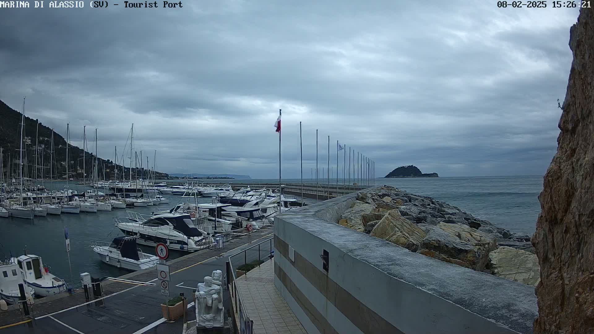 A harbor filled with yachts and sailboats is situated under an overcast sky, with a rocky breakwater separating it from a calm sea.