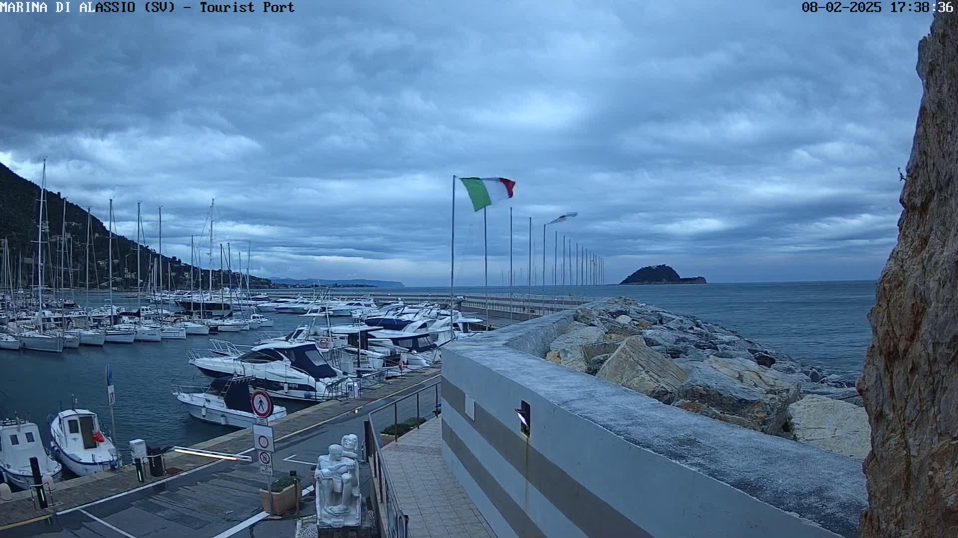 A harbor filled with sailboats and motorboats is seen under a cloudy sky, with a rocky breakwater and a small island visible in the distance.