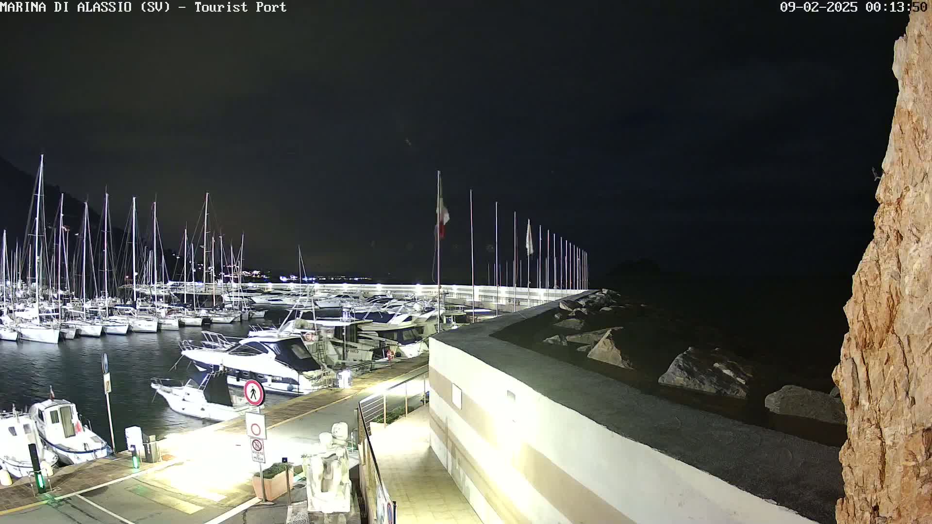 A nighttime view of a marina filled with sailboats and motorboats docked alongside a seawall, under a dark, clear sky.
