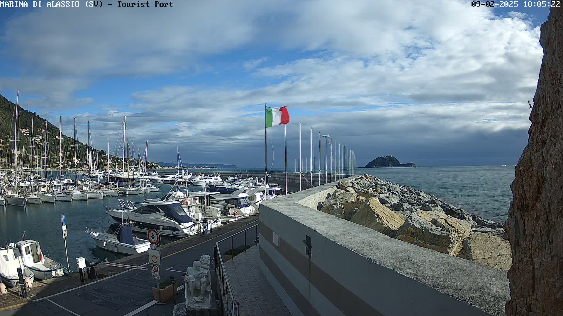 A harbor filled with numerous boats and yachts is situated next to a rocky breakwater under a partly cloudy sky.