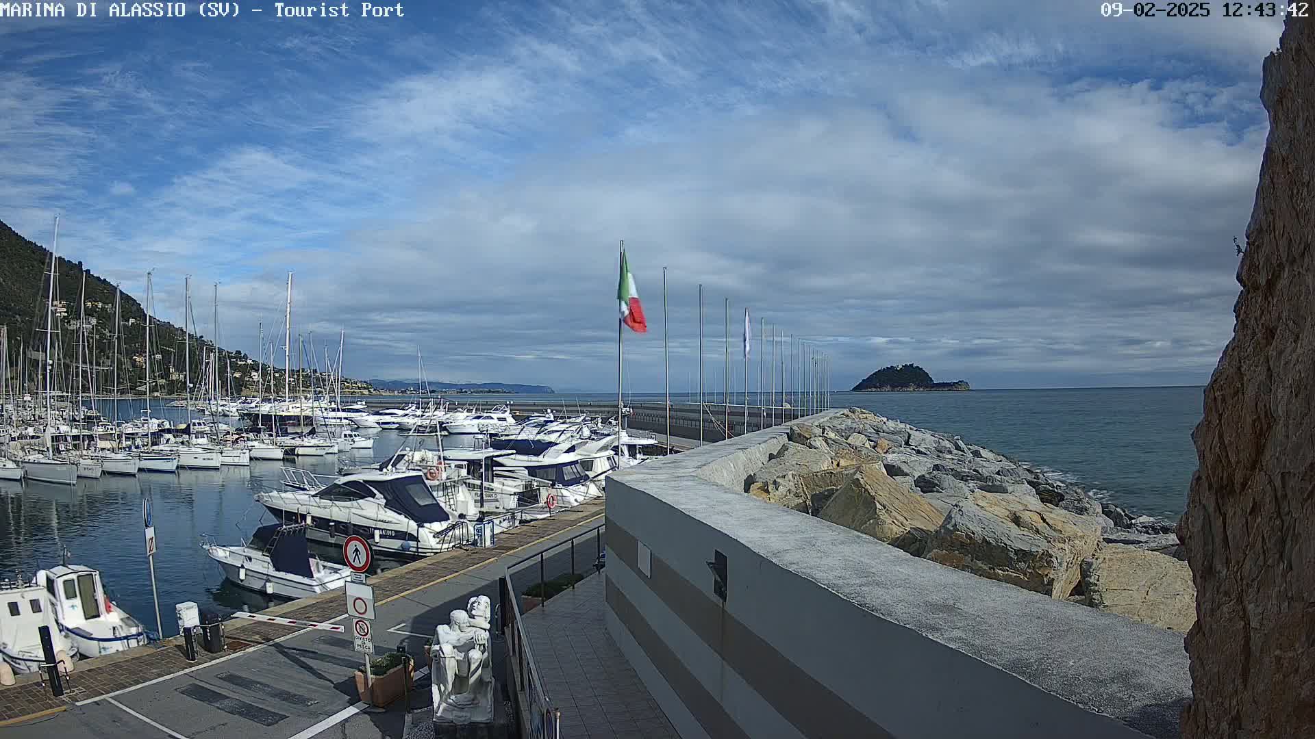 A marina filled with boats is situated next to a rocky shoreline under a partly cloudy sky.