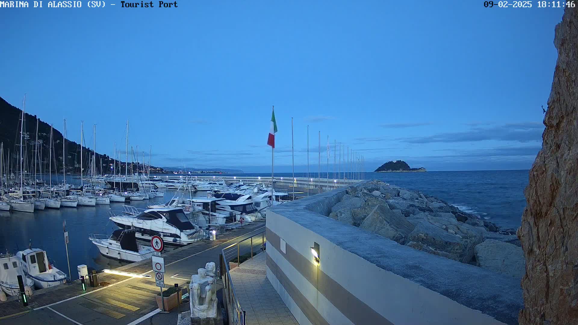 A marina filled with many boats is seen at dusk under a clear blue sky, with a rocky shore and a small island visible in the distance.