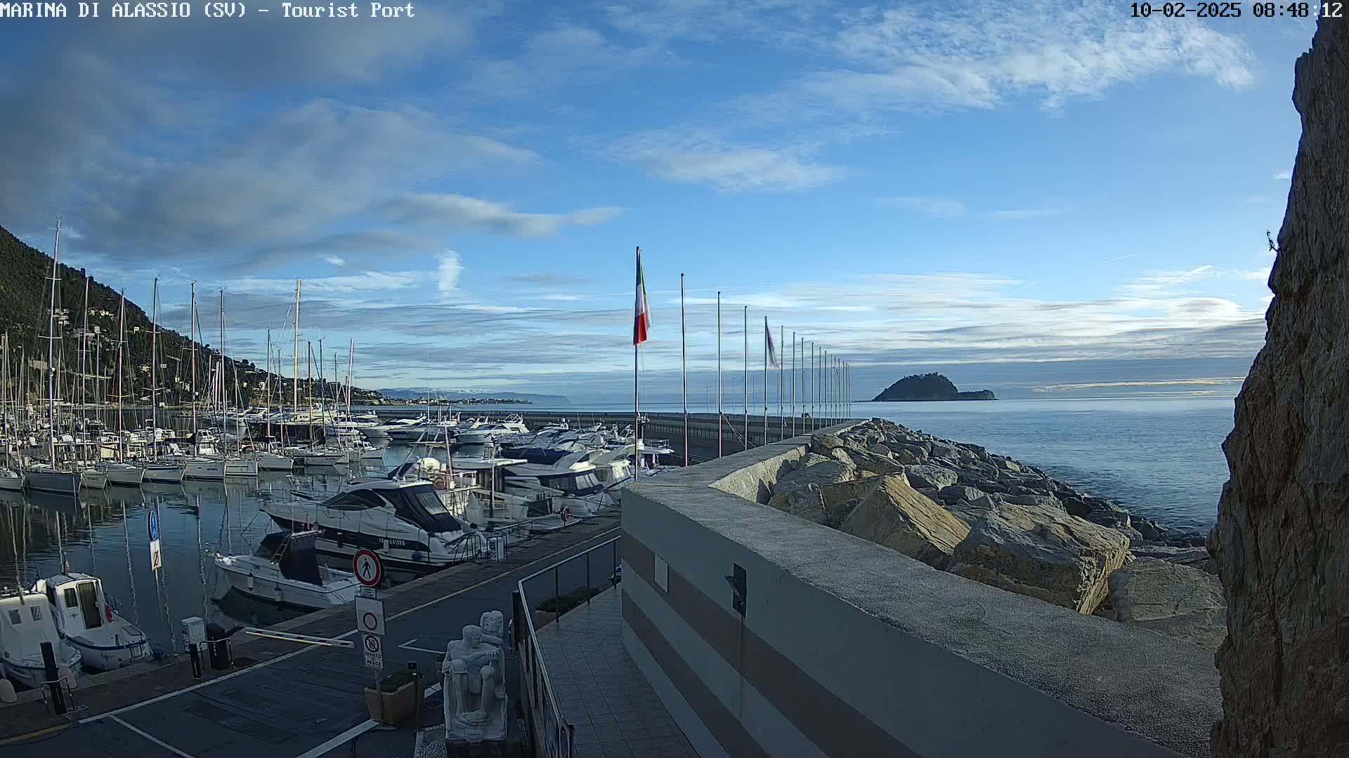 A marina filled with boats is situated on a calm sea under a mostly clear blue sky with some clouds.