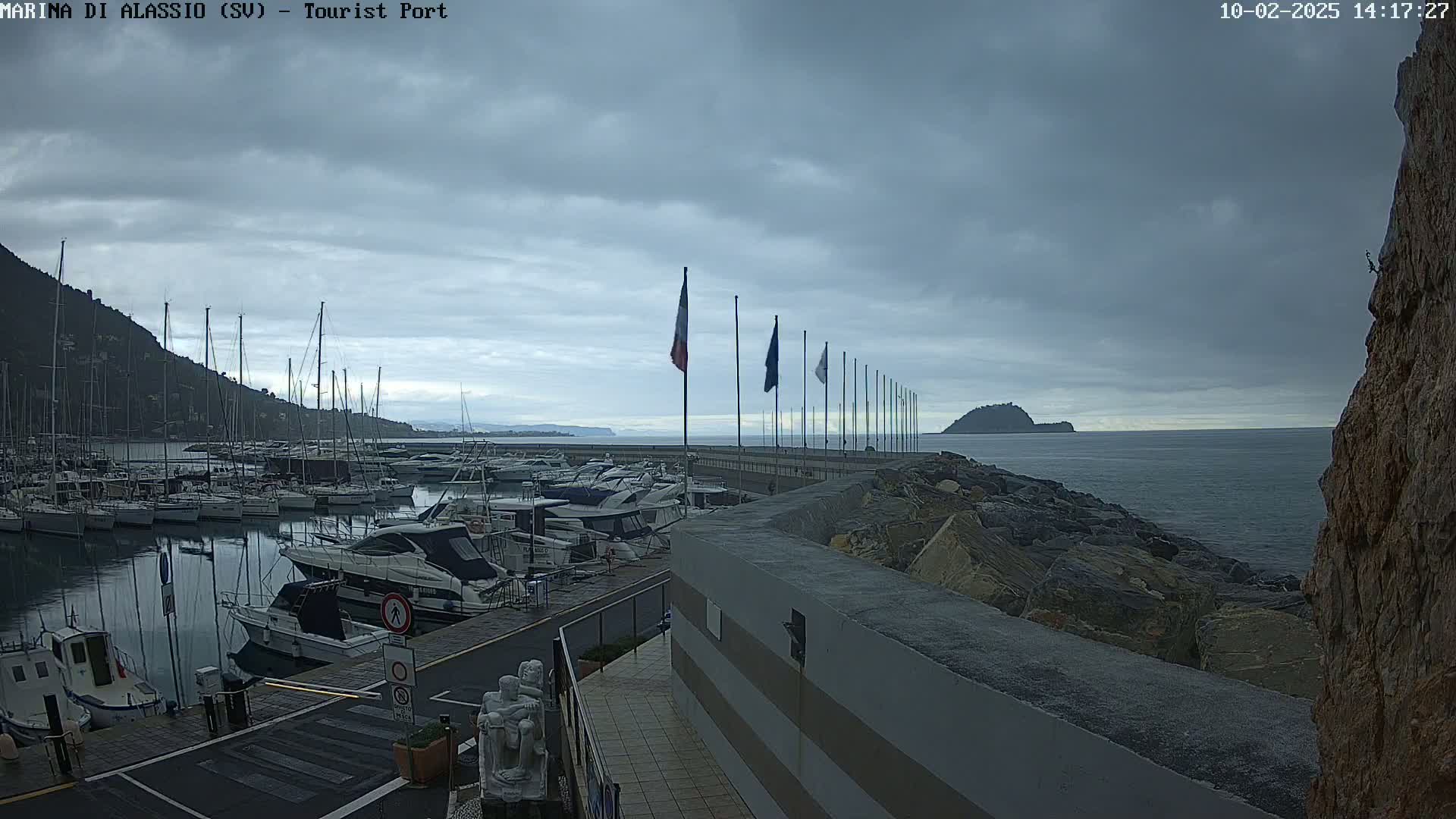 A harbor filled with sailboats and motorboats is seen under a cloudy sky, with a rocky shore and calm water extending to a distant island.