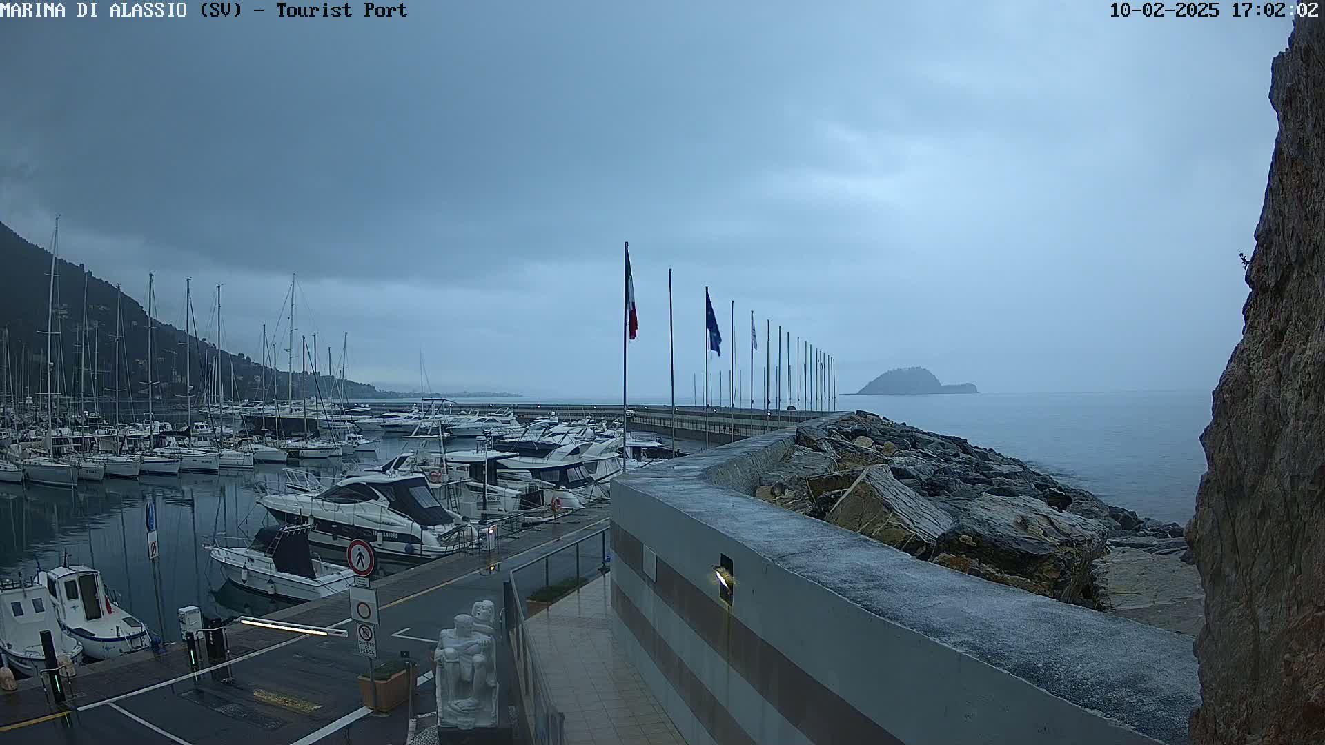 A harbor filled with boats is seen under an overcast sky, with a rocky shoreline and a small island in the distance.