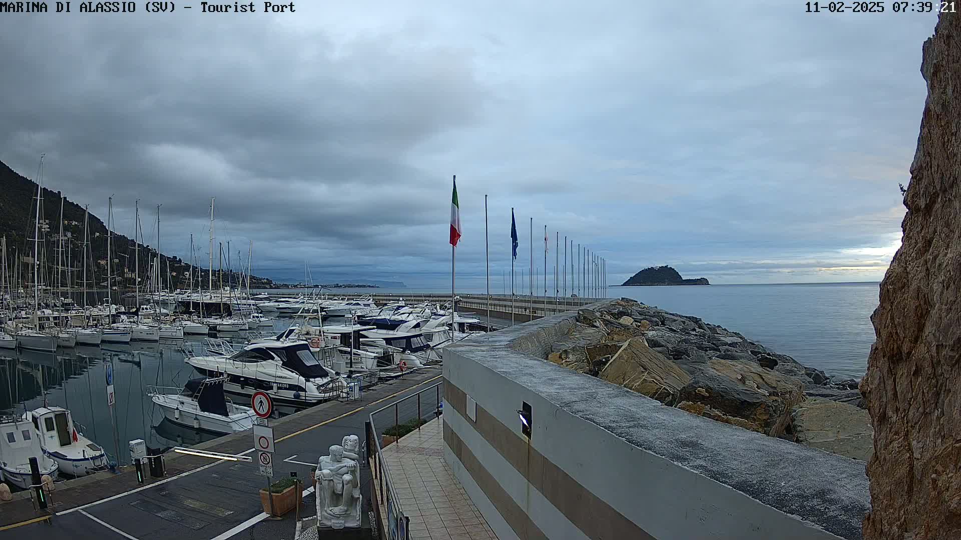 A marina filled with boats is situated next to a rocky shoreline under an overcast sky.