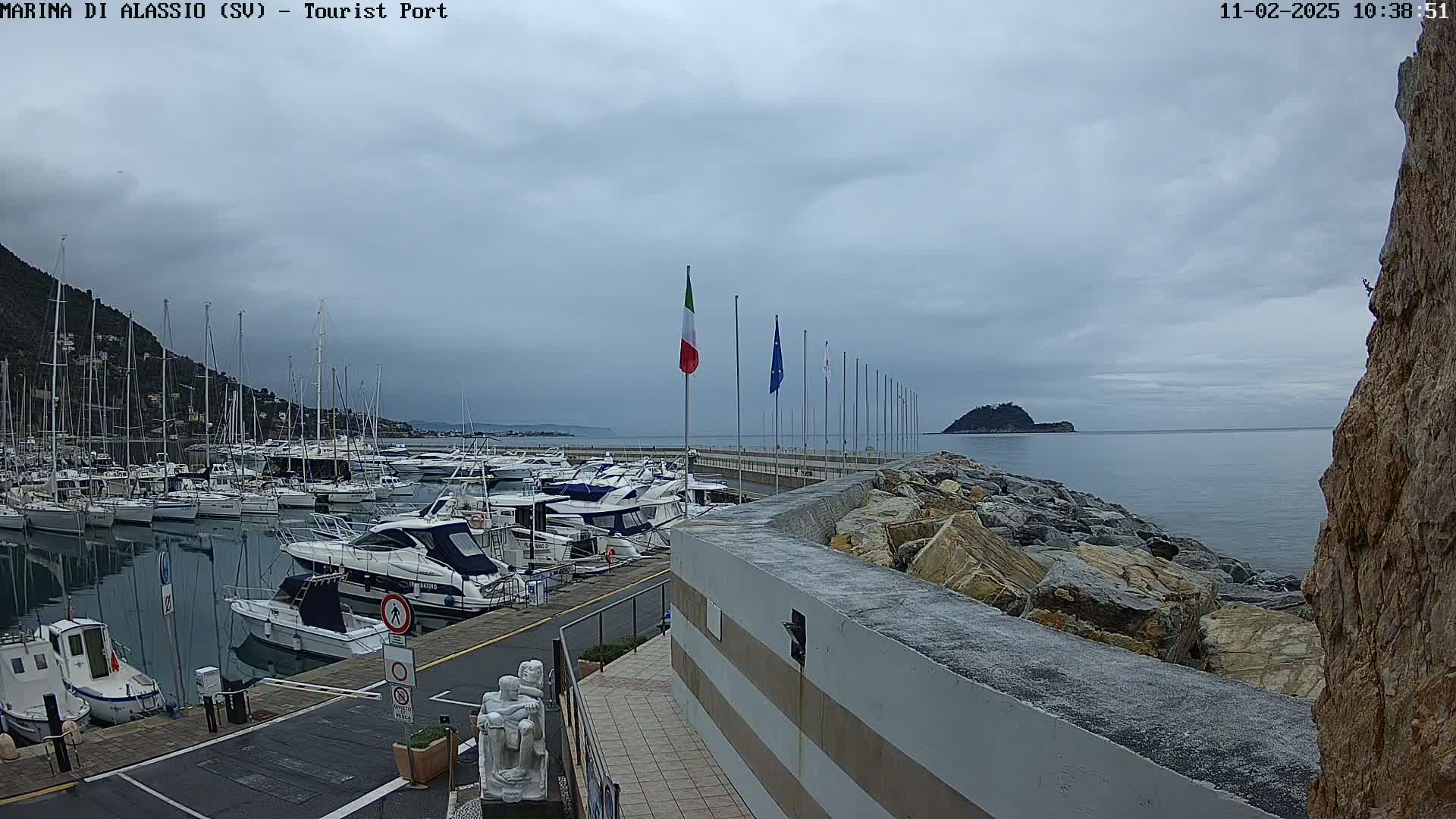 A marina filled with boats is situated on a calm sea under an overcast sky, with a rocky embankment and distant hills visible.