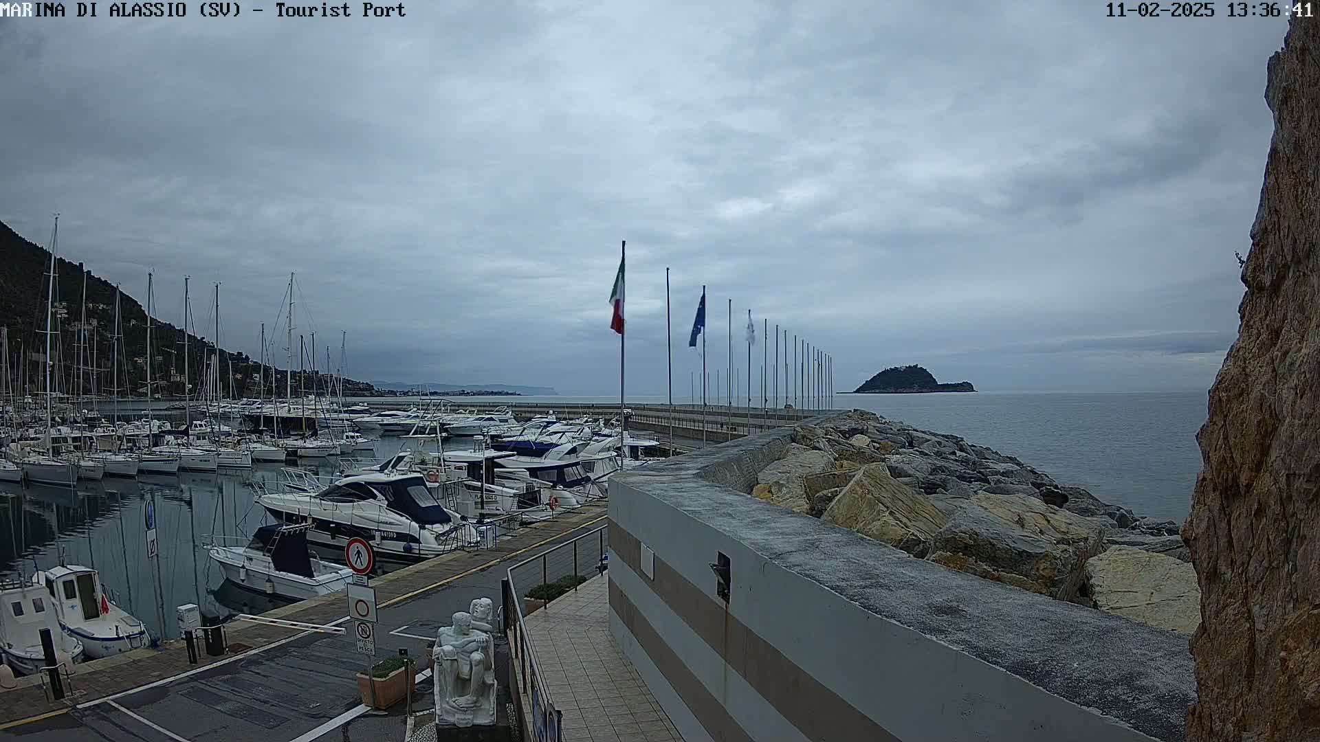 A marina filled with sailboats and motorboats is situated along a rocky coastline under a cloudy sky.