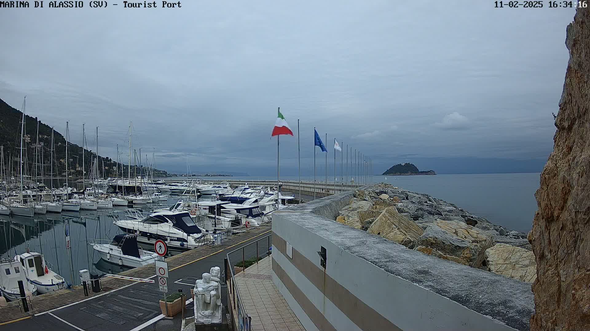 A marina filled with numerous yachts and sailboats is situated under a cloudy sky beside a rocky shoreline with Italian and European flags displayed.