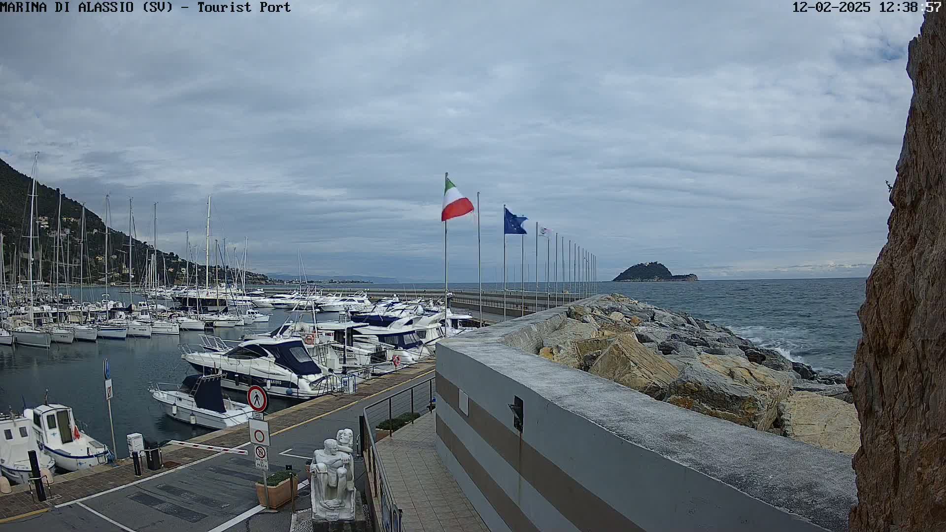 A harbor filled with numerous boats is situated next to a rocky shoreline under a cloudy sky, with small waves visible in the water.
