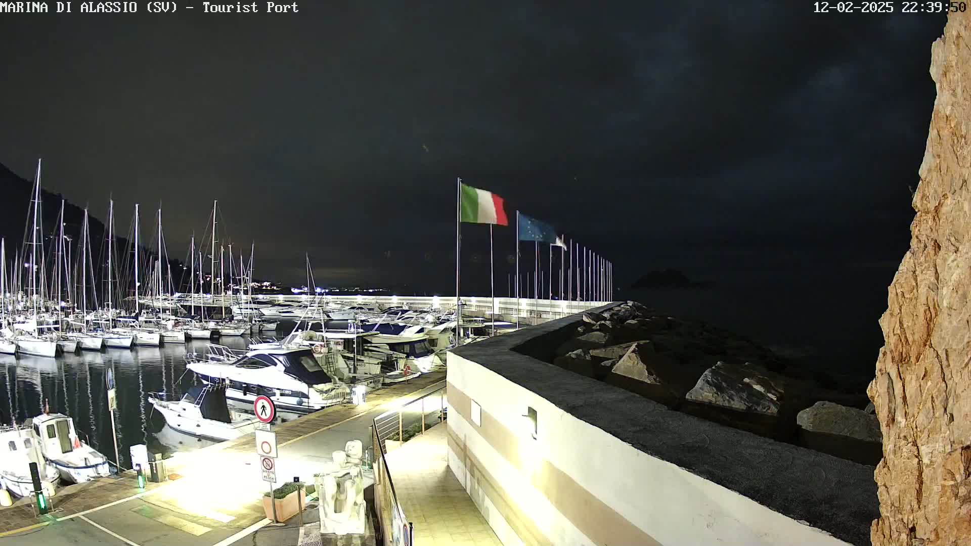 A nighttime view of a marina filled with boats, under a dark, cloudy sky, with Italian flags waving in the breeze.