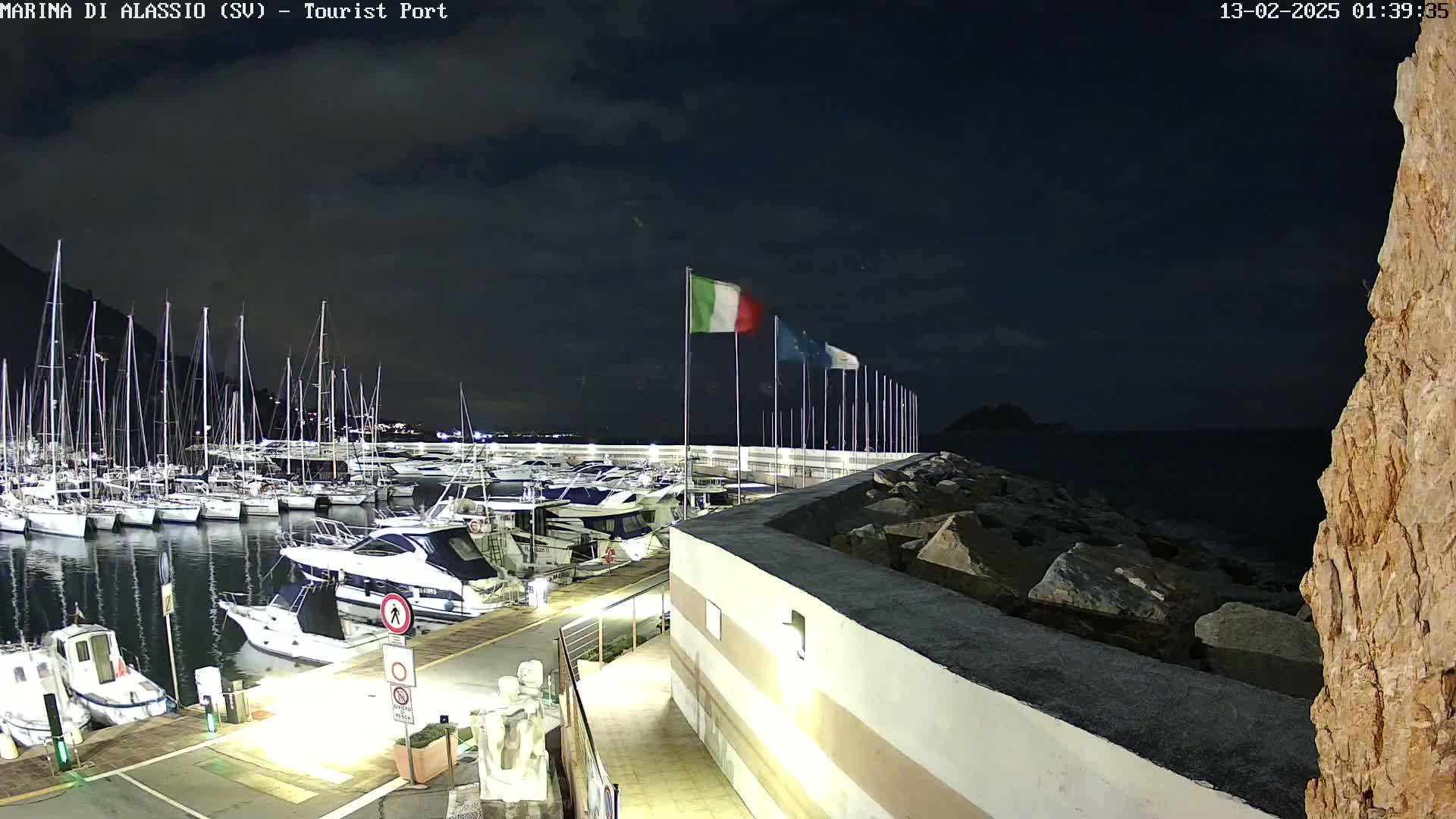 A nighttime view of a marina filled with sailboats and motorboats, under a dark, cloudy sky.