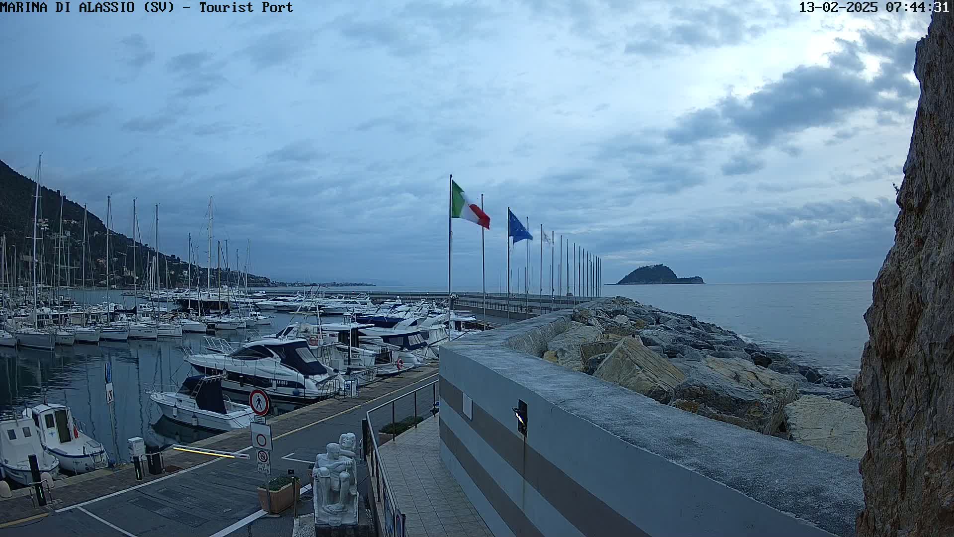A marina filled with numerous boats is situated near a rocky shoreline under a cloudy sky.