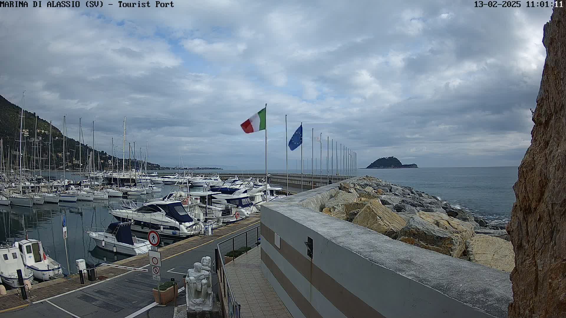 A harbor filled with numerous boats is situated next to a rocky shoreline under a cloudy sky.