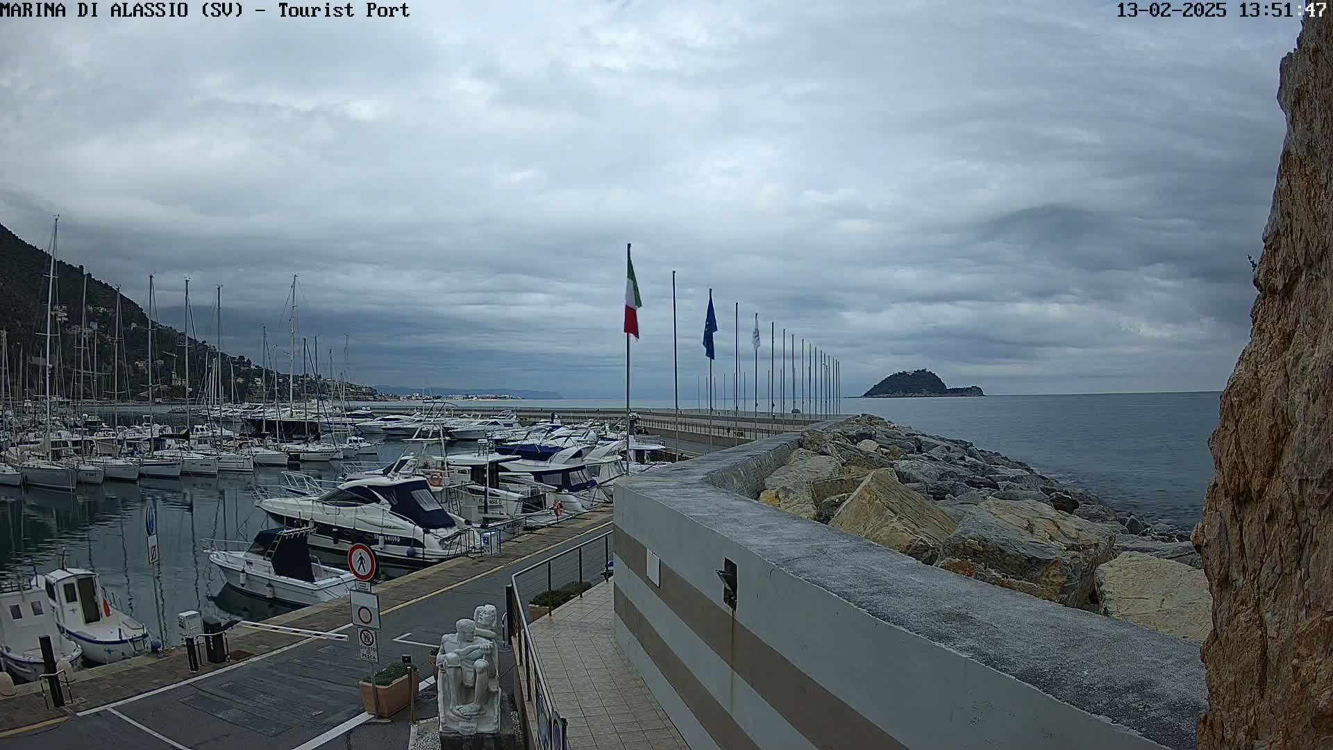 A harbor filled with boats is shown under a cloudy sky, with a rocky shoreline and a small island visible in the distance.