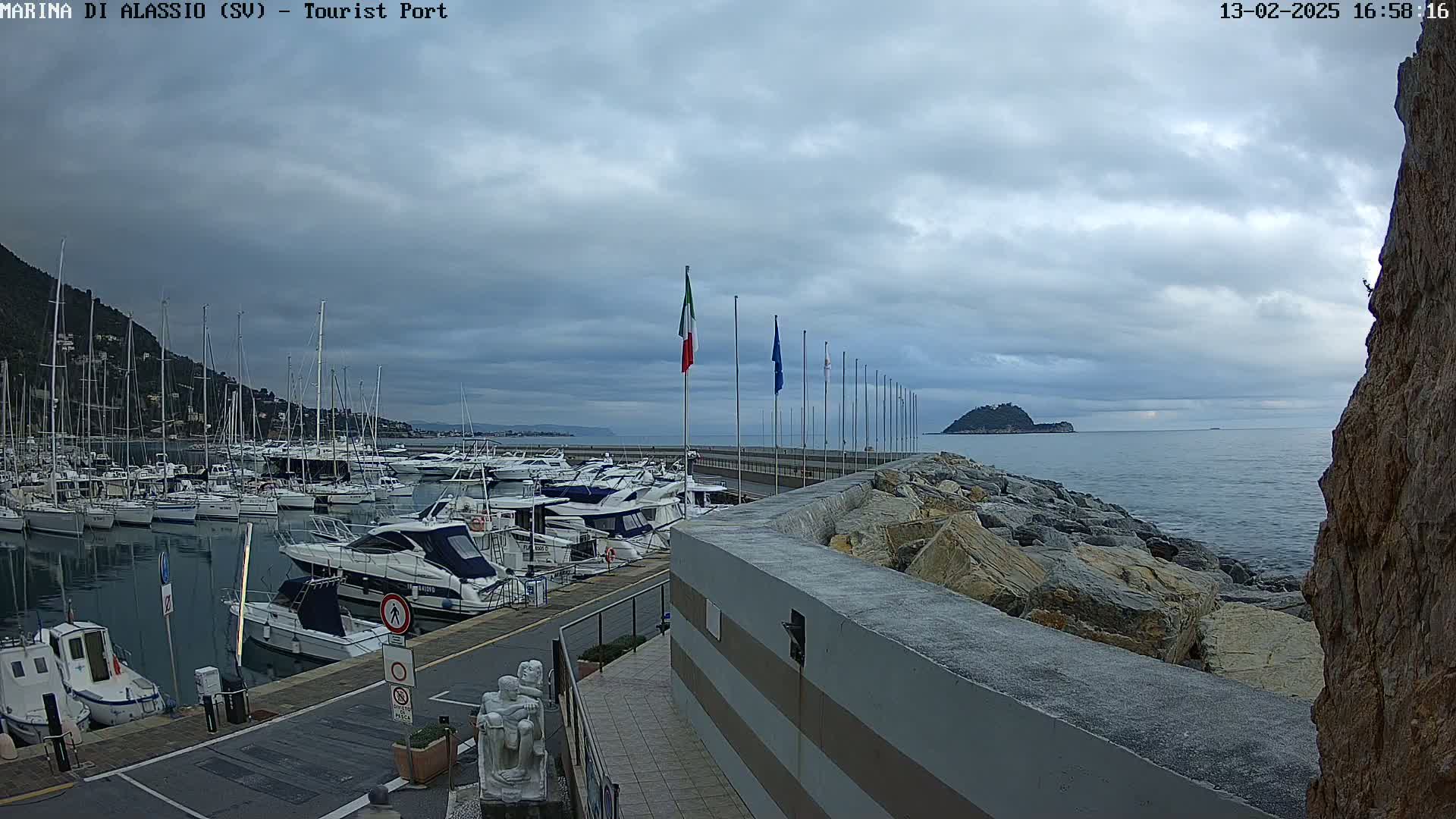 Many boats are docked in a harbor on a cloudy day, with a small island visible in the distance.