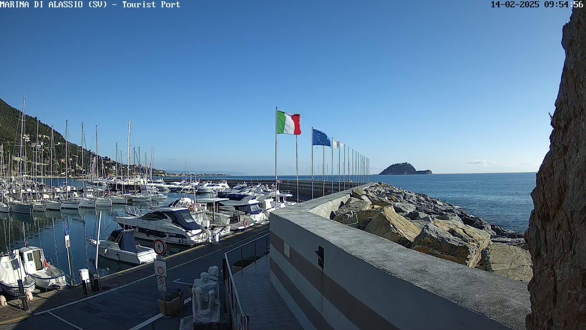 A marina filled with boats is situated on a calm, sunny day next to a rocky shoreline and a small island in the distance.