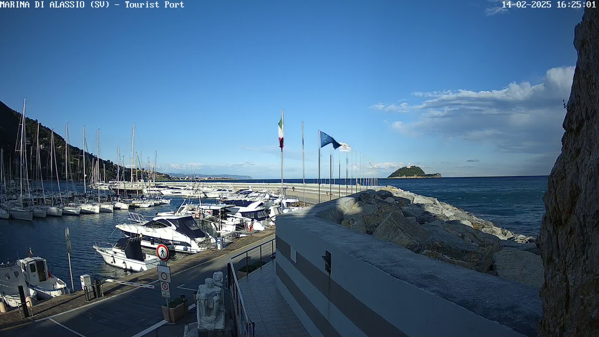 A marina filled with sailboats and motorboats is situated next to a rocky shoreline on a partly cloudy day.