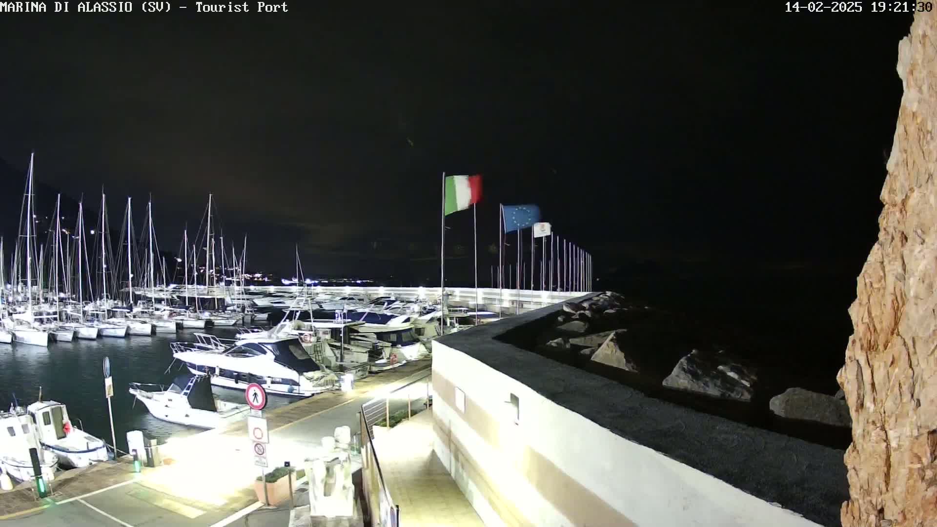 A nighttime view of a marina filled with sailboats and motorboats, under a dark sky with Italian and European Union flags waving.